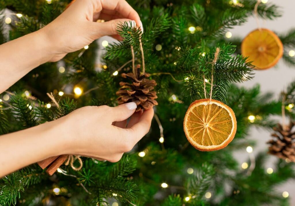 A woman hanging pinecones and orange slices as decorations on a tree.